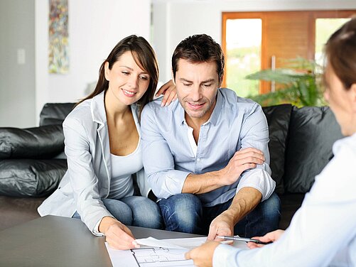 Ein Paar sitzt auf einem Sofa und schaut auf einen Plan, den eine dritte Person in der Nähe präsentiert. Die Frau lächelt, während der Mann aufmerksam den Plan betrachtet. Die Umgebung wirkt modern und freundlich.